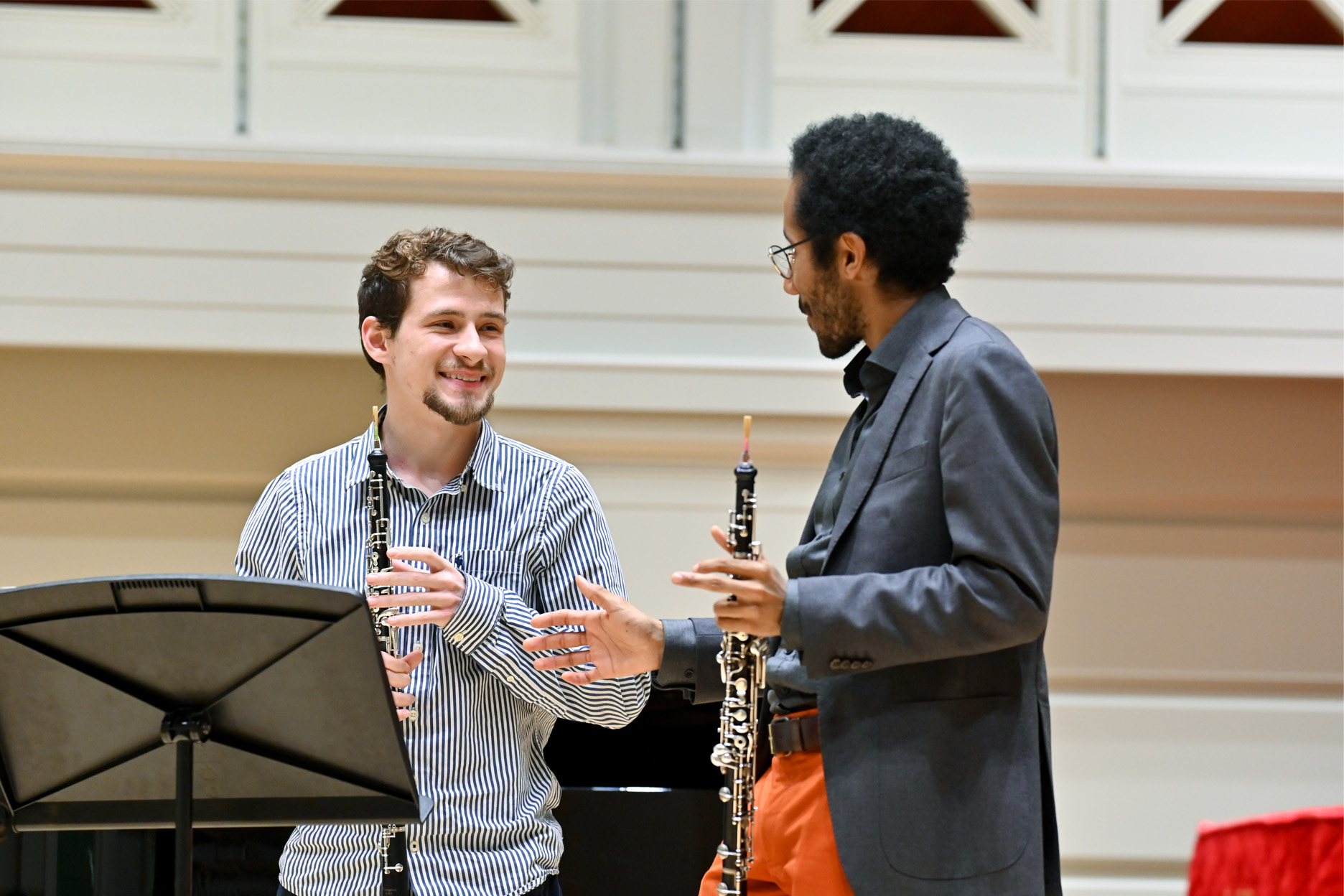 A male student holding his oboe with Armand Dijkoloum a man wearing a grey blue blazar, holding his oboe and smiling at him, while teaching.